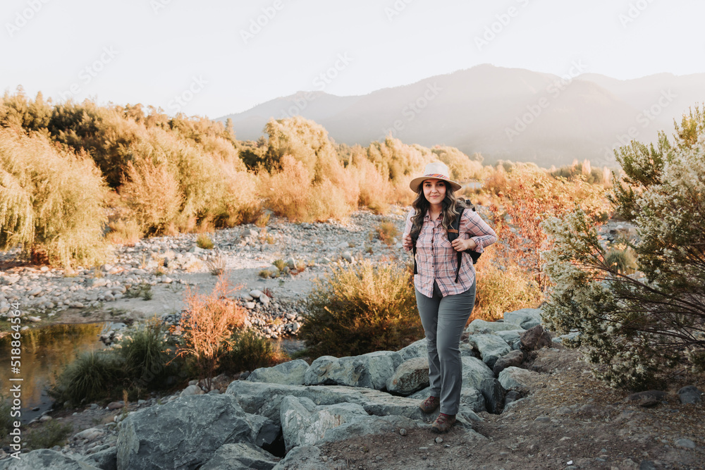 Smiling young latin woman with a hat and a backpack on, hiking in a beautiful landscape.