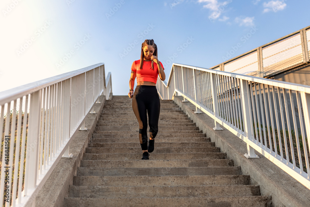 Young happy focused fitness girl in black yoga pants and orange short shirt runs downwards the bridge stairs during the day. Front view.
