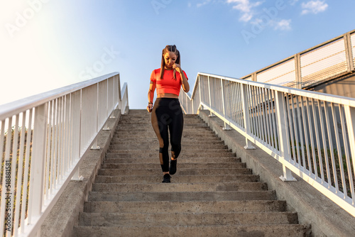 Young happy focused fitness girl in black yoga pants and orange short shirt runs downwards the bridge stairs during the day. Front view.