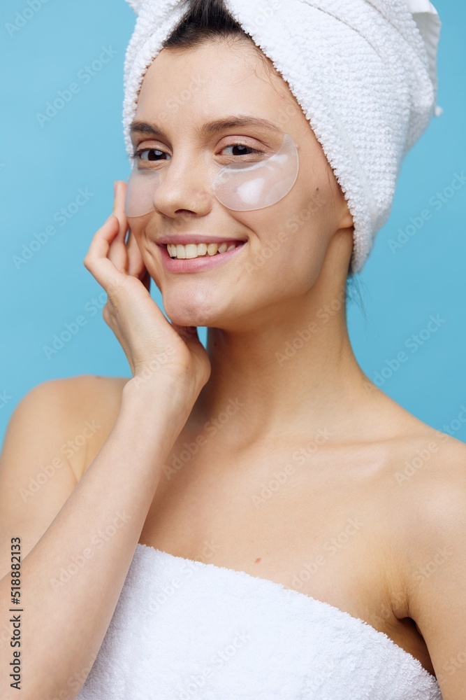 a beautiful woman stands on a blue background, with a towel on her head after a shower and patches pasted on her face, looks away holding her hand near her face and smiles happily a beautiful woman stands on a blue background, with a towel on her head after a shower and patches pasted on her face, looks away holding her hand near her face and smiles happily