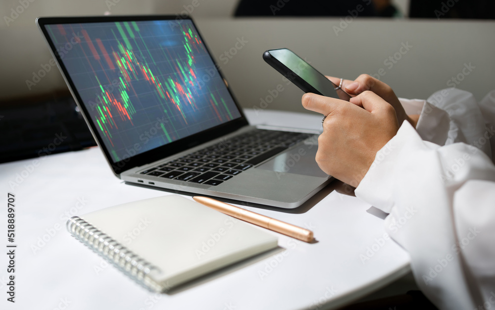 Young asian businesswoman using mobile phone to find information for investment analysis and analyze stock graph on laptop screen to make profitable plans. Close up view Young asian businesswoman using mobile phone to find information for investment analysis and analyze stock graph on laptop screen to make profitable plans. Close up view