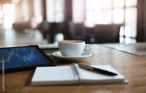 Close up view, tablet computer with graph on screen and white coffee cup on wooden table with note book and pen, concept business people relax and work part time in cafe, blurred and bokeh background