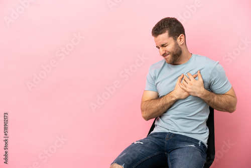 Young man sitting on a chair over isolated pink background having a pain in the heart © luismolinero