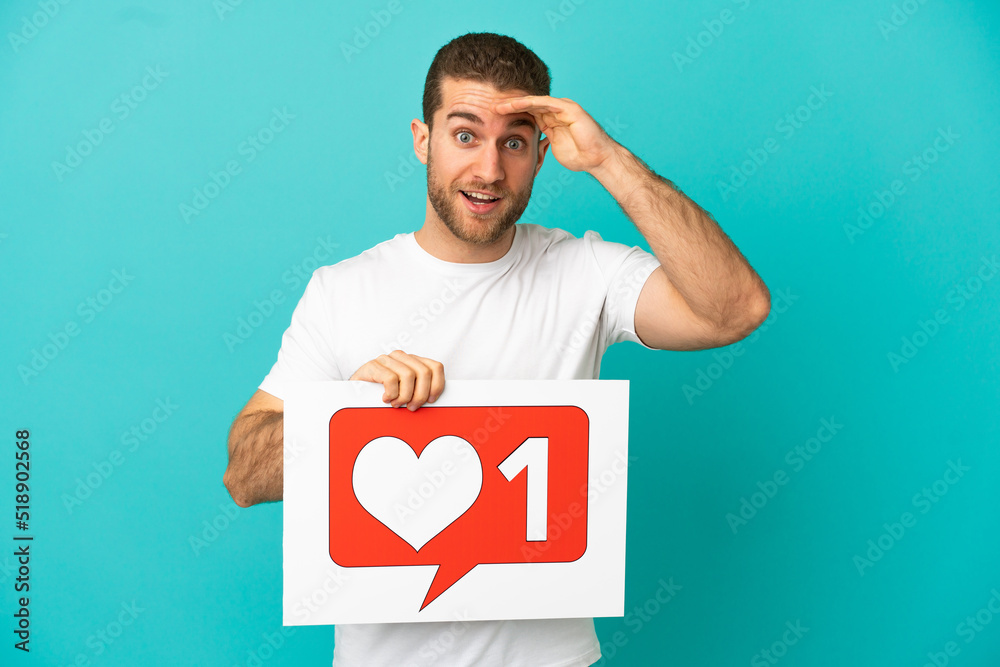 Handsome blonde man over isolated blue background holding a placard with Like icon with surprised expression Handsome blonde man over isolated blue background holding a placard with Like icon with surprised expression
