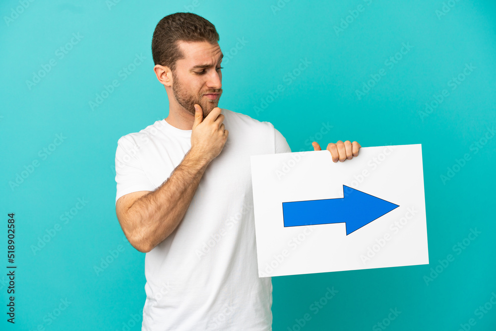 Handsome blonde man over isolated blue background holding a placard with arrow symbol and thinking Handsome blonde man over isolated blue background holding a placard with arrow symbol and thinking