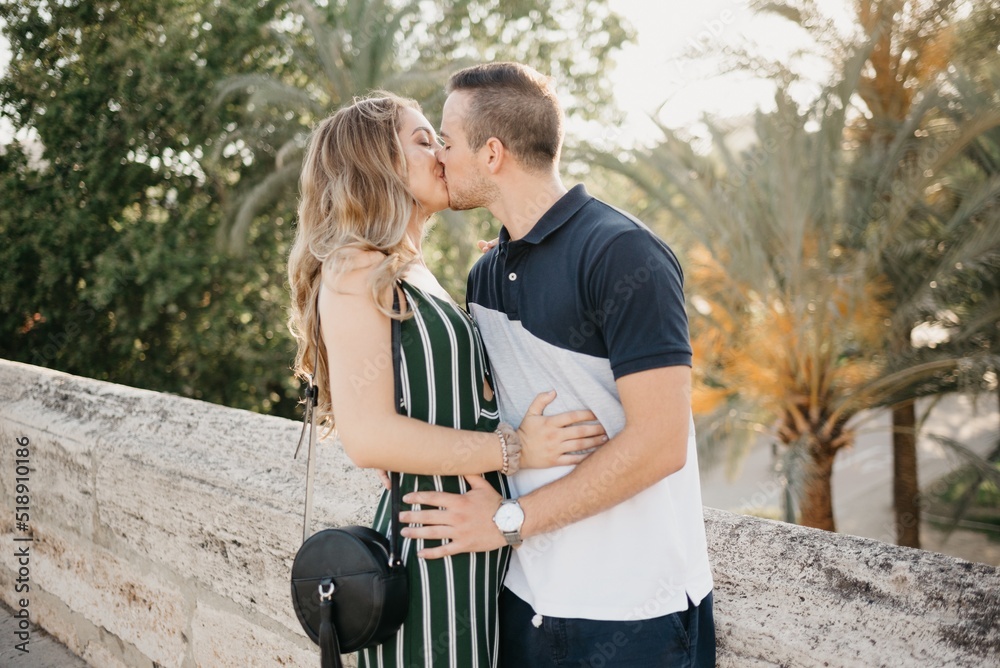 A girl in a green dress is kissing her boyfriend on the ancient bridge with palm trees in the background at the sunset in old Spain town. A couple of tourists on a date in the evening in Valencia.