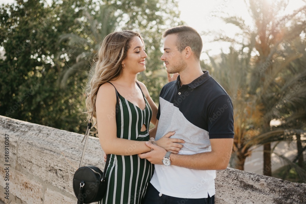 A girl in a green dress is hugging her boyfriend on the ancient bridge with palm trees in the background at the sunset in old Spain town. A couple of tourists on a date in the evening in Valencia.