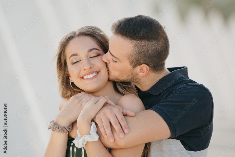 A close photo of a romantic guy who is kissing the cheek of his girlfriend in the modern urban space at the sunset in a Spain town. A couple of tourists on a date in the evening in Valencia.