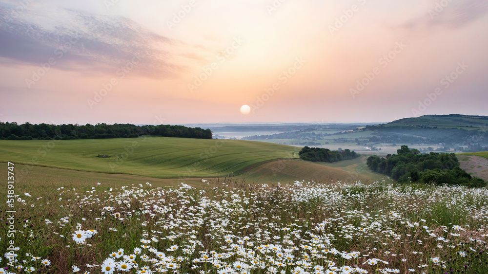 Beautiful English Summer sunrise landscape image looking over rolling hills with mist in distance and hazy sun low in the sky
