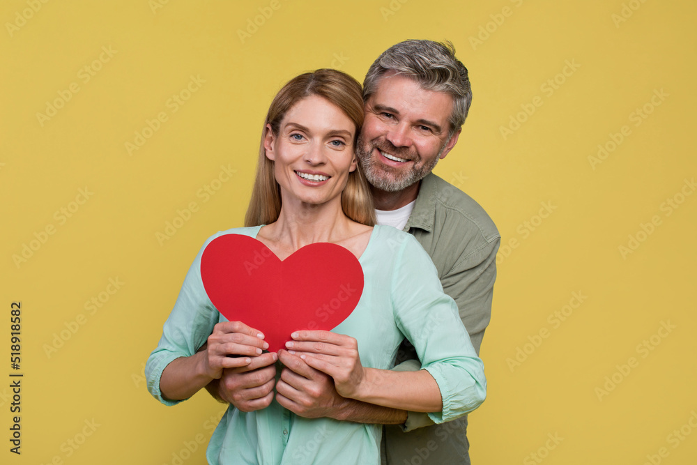 Loving middle aged spouses holding paper heart, man embracing wife brom back and smiling together at camera