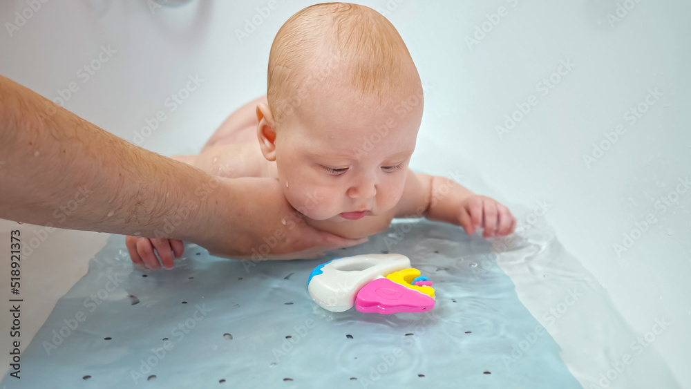 Father holds small daughter under belly in water. Baby girl plays with bright toy in bathtub. Child waves hands in water with amusement and curiosity Father holds small daughter under belly in water. Baby girl plays with bright toy in bathtub. Child waves hands in water with amusement and curiosity