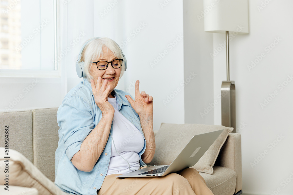 a happy elderly lady with gray hair is sitting on a cozy sofa talking via video link via laptop and headphones and smiling broadly holding her hands in a relaxed gesture a happy elderly lady with gray hair is sitting on a cozy sofa talking via video link via laptop and headphones and smiling broadly holding her hands in a relaxed gesture