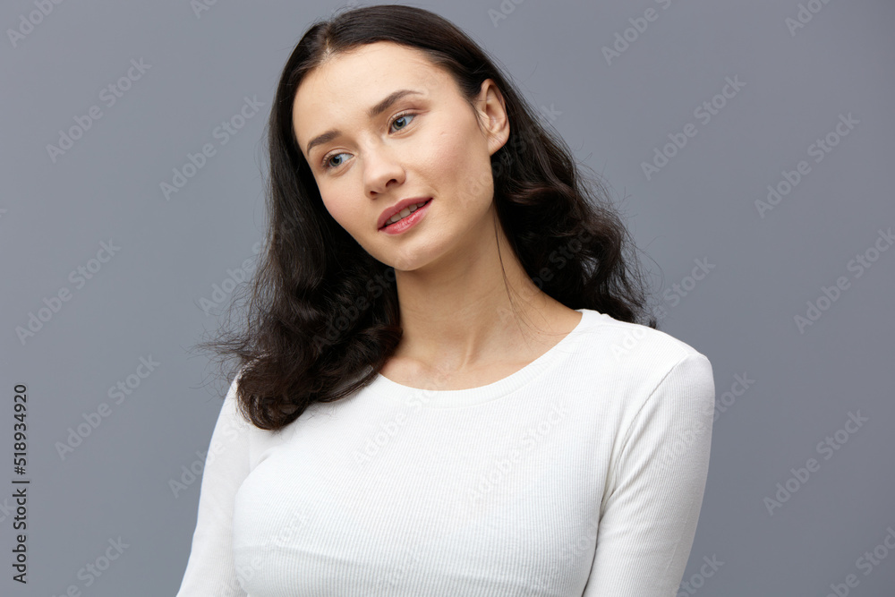 an attractive woman is standing on a gray background in a white T-shirt, with her hands behind her back, turning her head in different directions in a relaxed manner an attractive woman is standing on a gray background in a white T-shirt, with her hands behind her back, turning her head in different directions in a relaxed manner