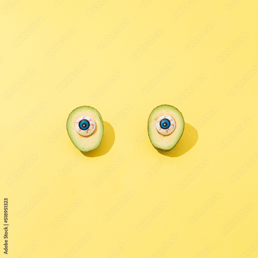 Sliced avocado with plastic eyes instead of stone on yellow background. Happy halloween concept with copy space.  Square ratio.