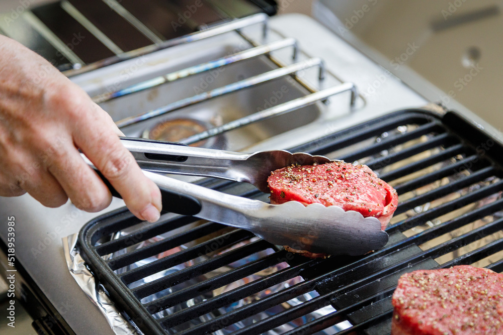 Using tongs to place bacon wrapped filet mignon steak on outdoor camping  grill Photos | Adobe Stock