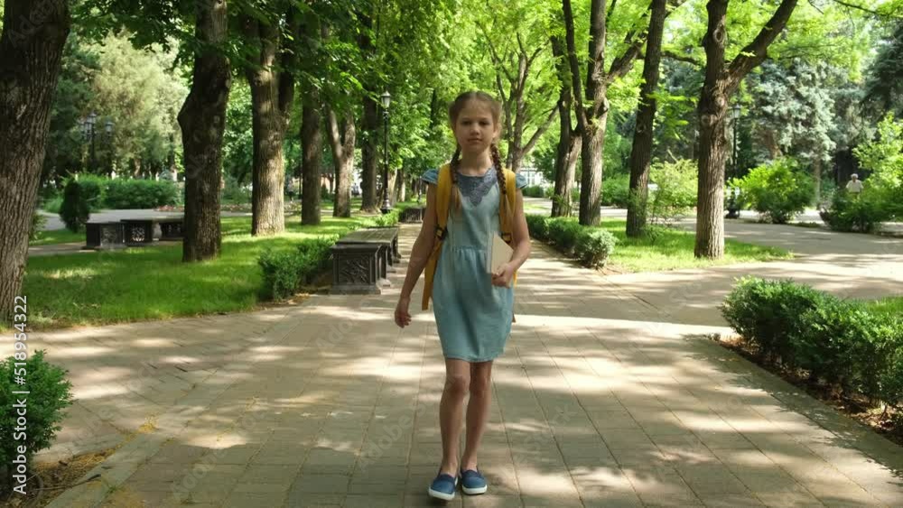 Back to school, education. School girl walks through the park alley to class in the morning. Little girl pupil goes after school with backpack and book. Beginning of lessons. First day of classes Back to school, education. School girl walks through the park alley to class in the morning. Little girl pupil goes after school with backpack and book. Beginning of lessons. First day of classes