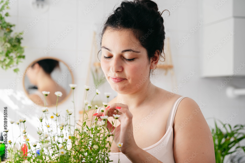 Young overweigh woman looking at chamomile flowers in white bathroom. Natural cosmetics concept. Body posititvity Young overweigh woman looking at chamomile flowers in white bathroom. Natural cosmetics concept. Body posititvity