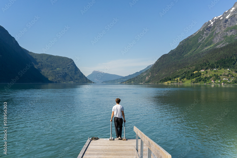 person standing at end of pier gazing at blue sky, mountains and water