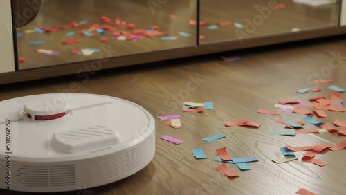 The robot vacuum cleaner does the cleaning in the apartment. A vacuum cleaner cleans up colorful candies after a holiday in a house with a brown wooden floor and a mirror. Smart House