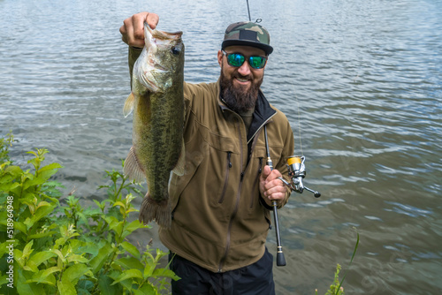 Bass fishing. Large bass fish in hands of pleased bearded fisherman with tackle. Largemouth perch at pond