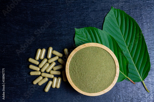 Mitragyna Speciosa Korth or kratom powder on wooden bowl wtih capsules and green leaf on black wooden background, top view and copy space.  photo