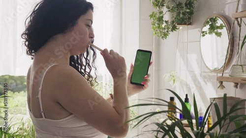 Young beautiful woman with overweight brushing her teeth and looking at the phone in light batroom with many green plants. Wellness, everyday technology. 4K UHD