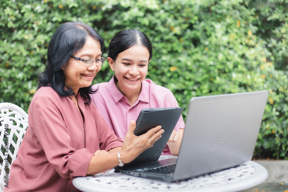 Happy Asian woman teaching her mother to use tablet and laptop in home garden. Senior mature and young woman surfing internet with digital devices together. Middle aged and technology Happy Asian woman teaching her mother to use tablet and laptop in home garden. Senior mature and young woman surfing internet with digital devices together. Middle aged and technology