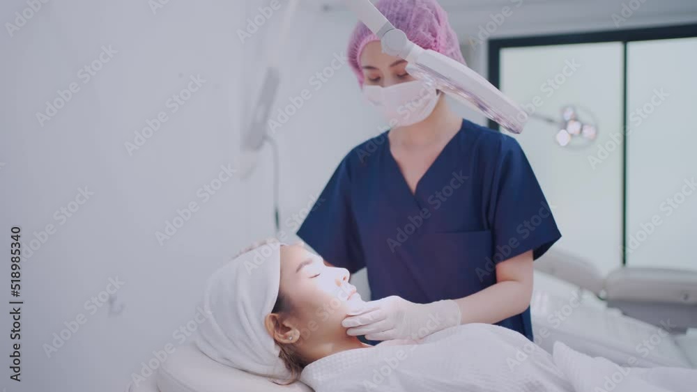 Young Asian female nurse in blue clothes, medical hat, face mask, and gloves moved LED light to shine and examine the face of Asian female client lying on bed in treatment room in a beauty clinic.