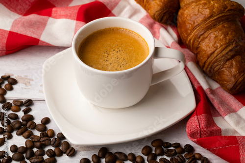 A cup of coffee with croissants and spilled coffee beans on a white wooden table.