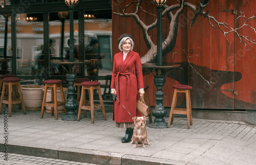 Elderly senior woman walking with lovely dog pet in red coat. Old retired aged lady on walk on city street with flowers.Stylish elder person pensioner. Quite calm happy years in metropolis © velirina