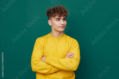 a handsome man stands on a green background in a yellow sweater and smiling pleasantly poses with his arms folded on his chest . Horizontal photo with empty space