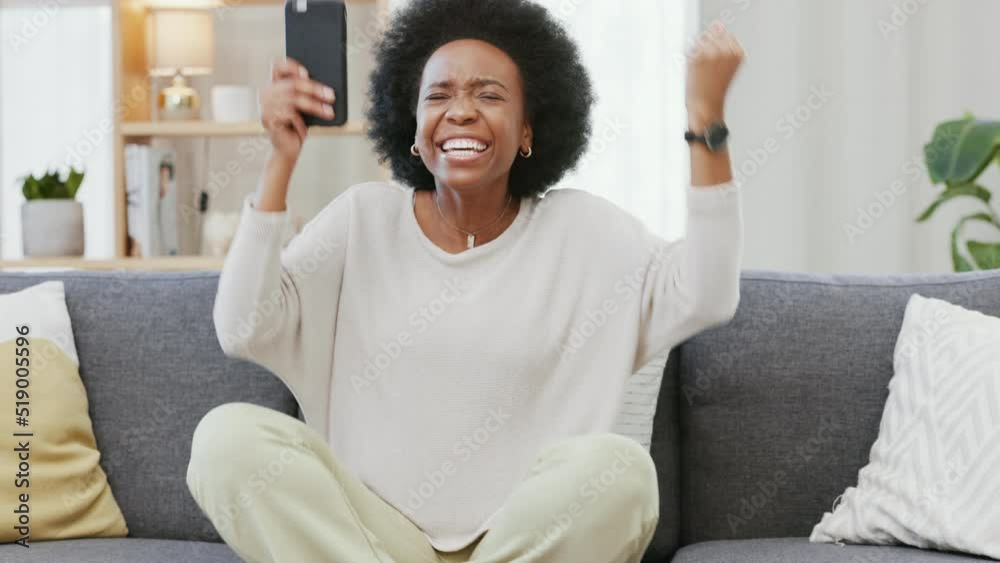 Happy African American woman expressing joy with a winning gesture while holding a phone and sitting on a sofa. Black female surprised by good news or results. Lady getting an approved business loan