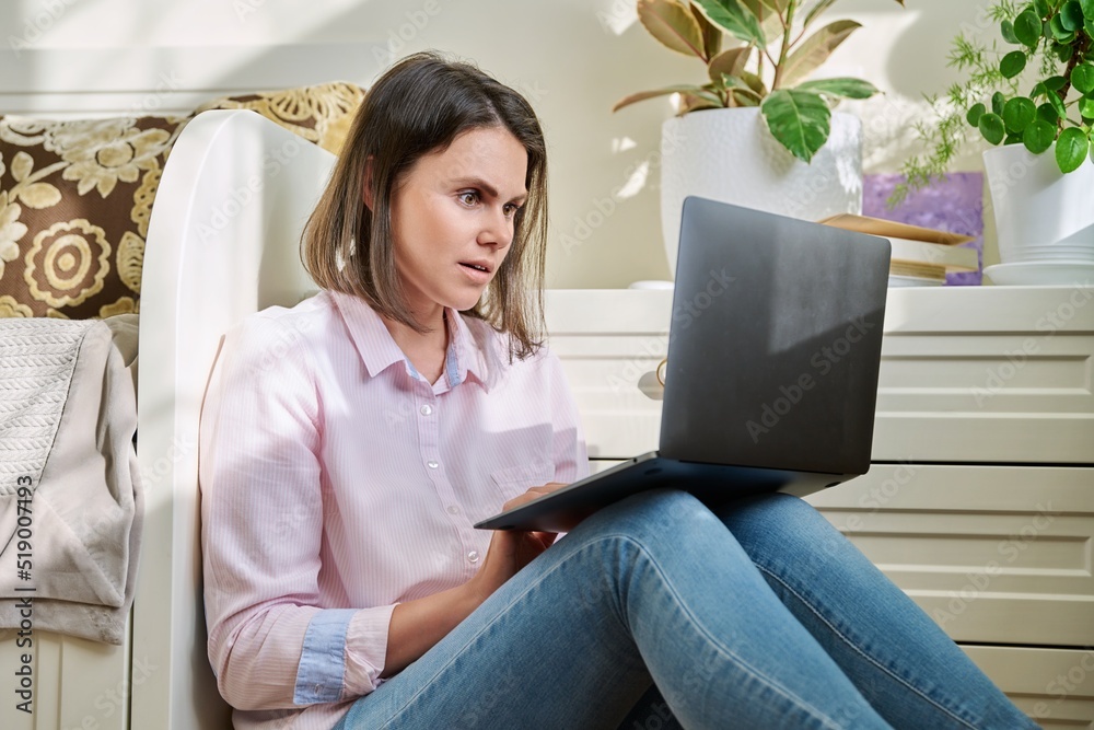 Serious sad young woman looking at laptop screen, sitting on floor at home Serious sad young woman looking at laptop screen, sitting on floor at home