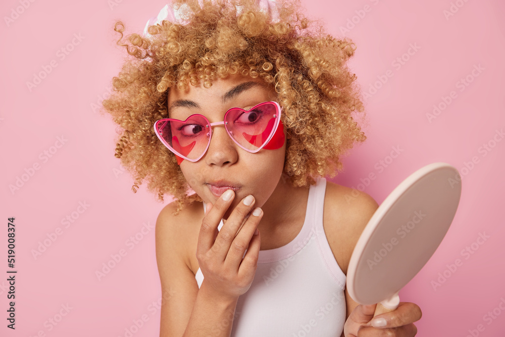 Indoor shot of serious curly haired woman applies hydrogel patches under eyes looks at herself in mirror wears heart shaped sunglasses and t shirt isolated over pink background. Beauty concept Indoor shot of serious curly haired woman applies hydrogel patches under eyes looks at herself in mirror wears heart shaped sunglasses and t shirt isolated over pink background. Beauty concept