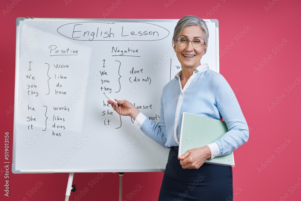Fototapeta premium Cheerful senior woman teaching English language near the whiteboard against pink background