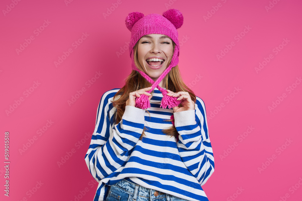 Playful young woman adjusting her pink hat and smiling while standing against colored background Playful young woman adjusting her pink hat and smiling while standing against colored background