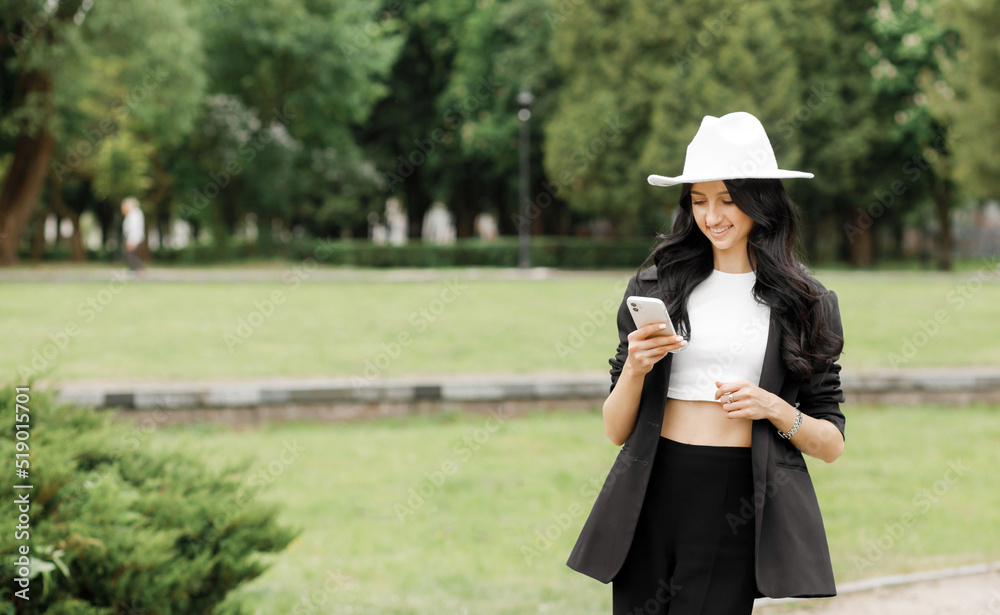 Business woman in a suit and hat walking along an park, checking her smartphone