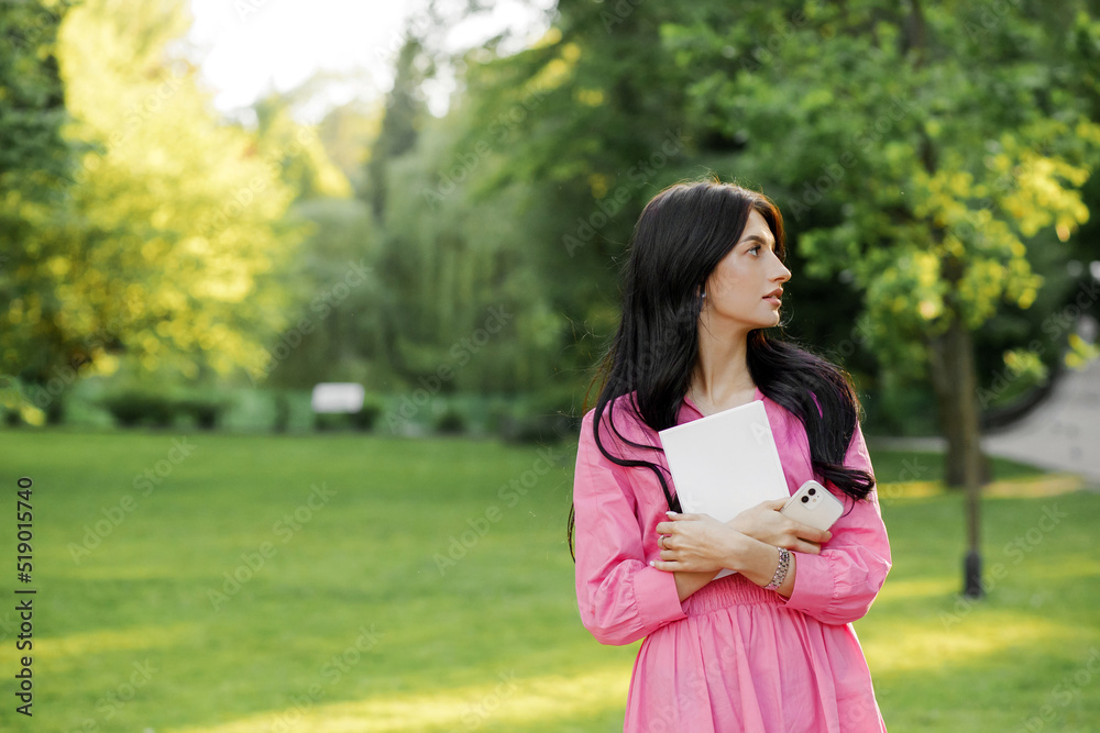 Close up portrait of young woman with wavy hair in pink dress in park holding book in hands. Learning concept Close up portrait of young woman with wavy hair in pink dress in park holding book in hands. Learning concept