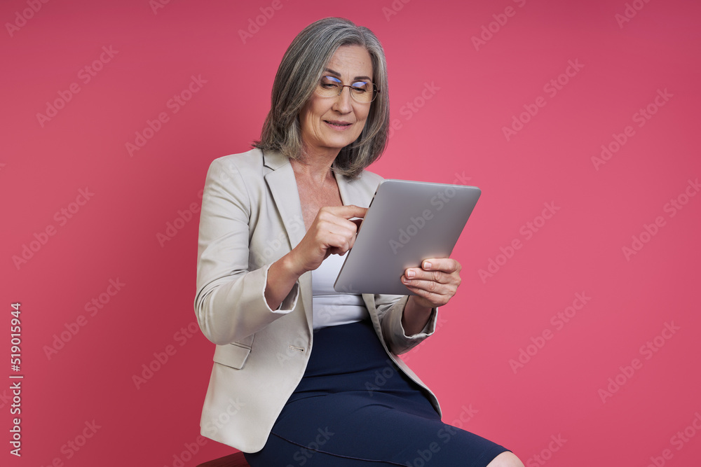 Confident senior woman in formalwear using digital tablet while sitting against pink background Confident senior woman in formalwear using digital tablet while sitting against pink background