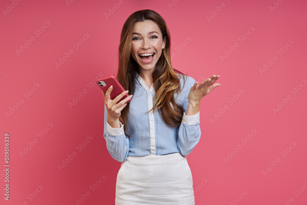 Happy young woman holding smart phone and gesturing while standing against colored background Happy young woman holding smart phone and gesturing while standing against colored background
