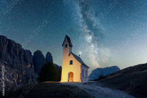 Incredible view on small iIlluminated chapel - Kapelle Ciapela on Gardena Pass, Italian Dolomites mountains. Glowing Milky Way in starry sky on background. Dolomite Alps, Italy. Landscape photography