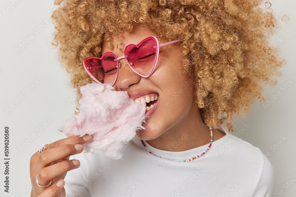 Φωτογραφία Close up shot of curly haired beautiful woman eats appetizing candy floss enjoys favorite dessert wears heart shaped sunglasses and t shirt isolated over white background