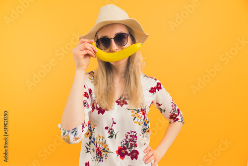 European woman in her 20s in summer hat, big sunglasses and floral blouse covering her mouth with a long yellow banana creating a smile. Orange background. Isolated copy space studio shot. High