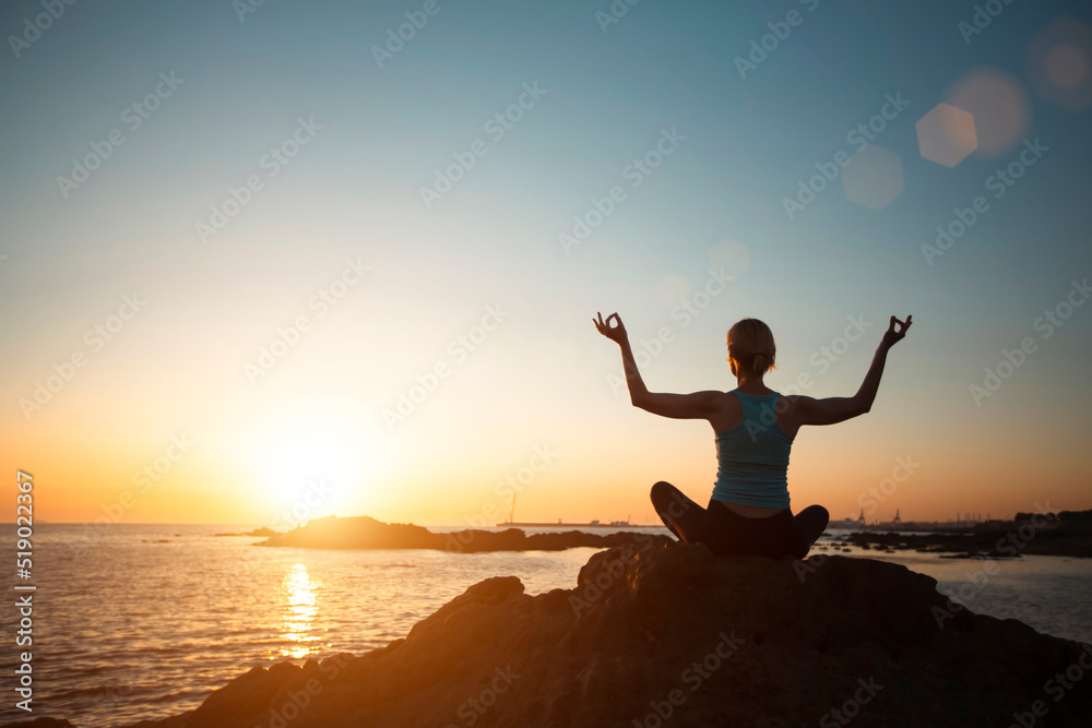 A woman does yoga in the lotus pose on the ocean, during sunset.