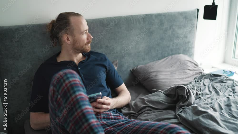 Meditative blond man looking at view and typing on phone while sitting on the bed at home Meditative blond man looking at view and typing on phone while sitting on the bed at home