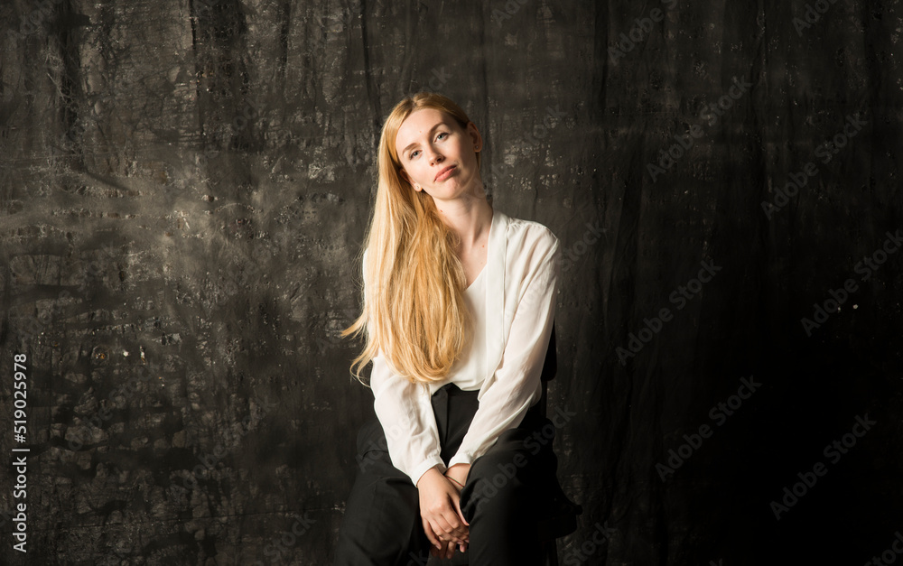 portrait of a beautiful blonde woman with long hair in a white blouse and black trousers on a gray isolated background.a charming girl posing on a dark background. portrait of a beautiful blonde woman with long hair in a white blouse and black trousers on a gray isolated background.a charming girl posing on a dark background.