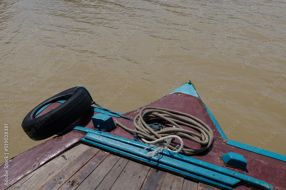 Fototapeta premium Low angle sunny view at the bow, front of the boat, with wooden floor float straight ahead along river with background of riverside of the countryside of Ayutthaya province in Thailand. 