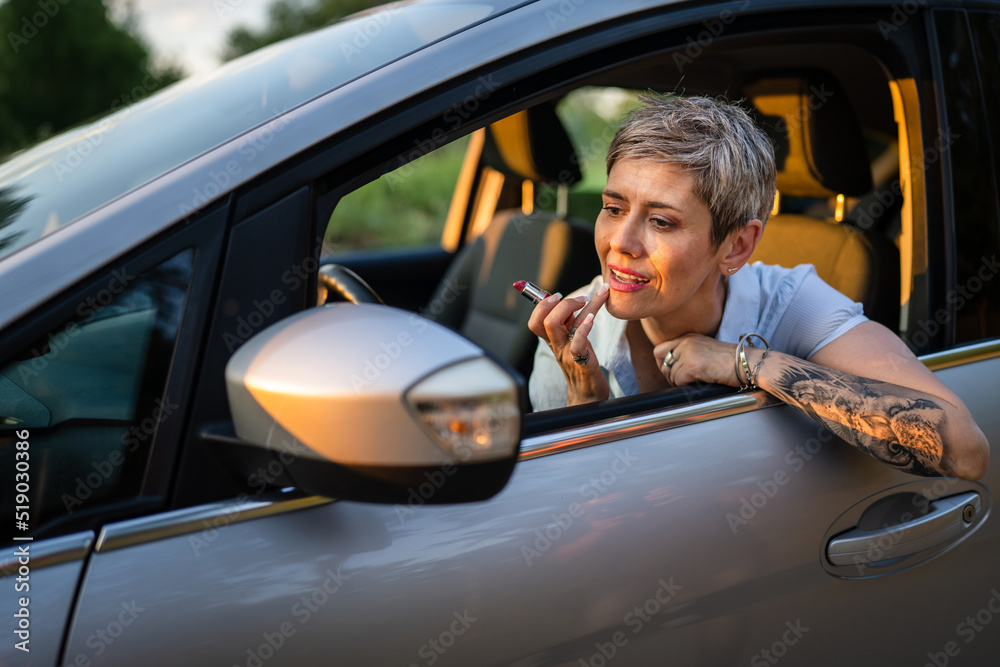 Naklejka premium One woman mature caucasian female businesswoman sitting in car putting lipstick fixing repairing makeup on her face while waiting in summer day evening real people copy space gray hair