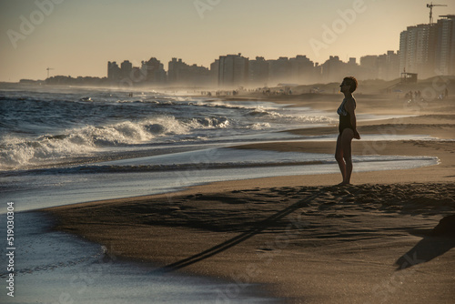 Mature woman standing in front of the sea looks at the waves that break, in the background the mist and buildings in Punta del Este, Uruguay. photo