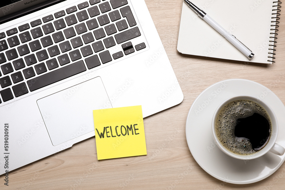 Paper note with word Welcome, laptop, coffee and notebook on wooden table, flat lay Paper note with word Welcome, laptop, coffee and notebook on wooden table, flat lay